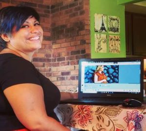 Sarah Perez, smilling, showing samples of her artwork on a laptop and hanging up.