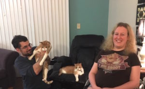 Katy Oliver, sitting in her wheelchair, with her husband holding one cat and another cat sitting in a chair. They are all in a living room. 
