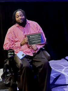 Edward Mitchell, a Black man with a short beard who uses a wheelchair, on a stage holding a plaque
