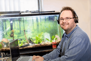 Ty Erd Smith, a white man with light, brown hair and a short beard, wearing headphones while seated with an open laptop in front of an aquarium.