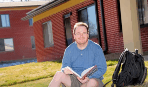 Ty Erd Smith, a white man with light, brown hair and a short beard seated on grass with a backpack and holding an open book near a brick building.