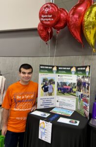 Emmanuel Alvarez, a young man wearing an orange Special Olympics Florida T-shirt stands beside an informational display for the Special Olympics Torch Icon Campaign at Publix Super Markets. The display features photographs of Special Olympics participants, messages about unity and red and gold balloons overhead, including one bearing the Special Olympics Florida logo.